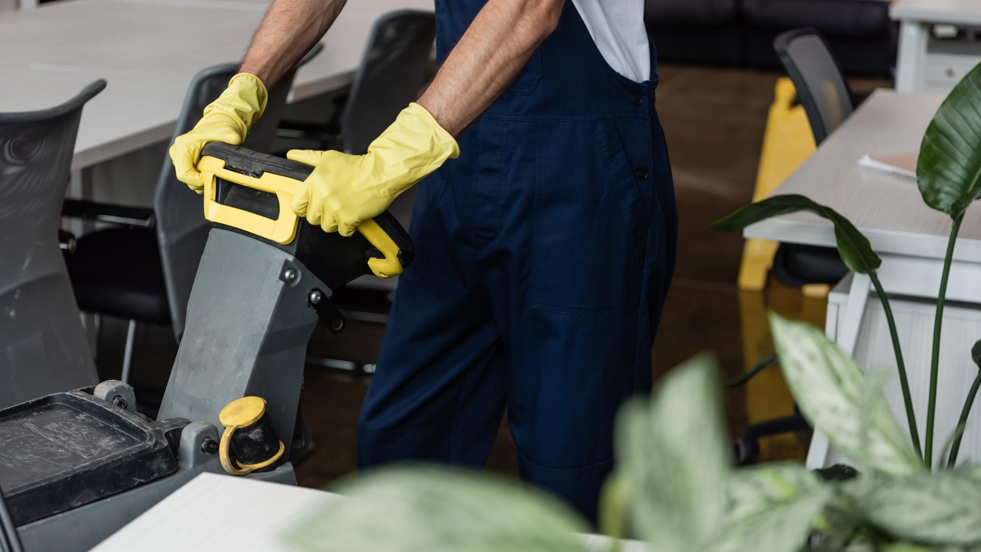A person wearing yellow rubber gloves operates a cleaning machine in an office setting, surrounded by desks and plants.